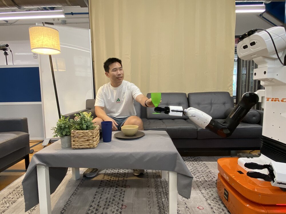 A person seated on a sofa holds a green cup above a TIAGo robot's outstretched gripper in a home-like lab setting.